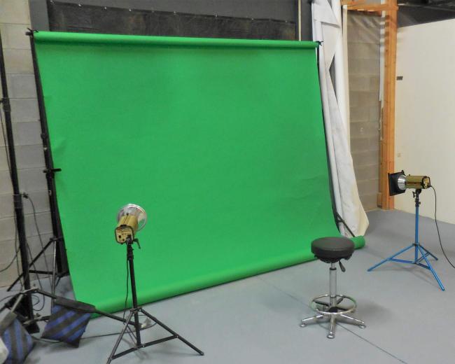 A studio setup with a green screen, two lights, a stool, and a partially visible workspace in the background.