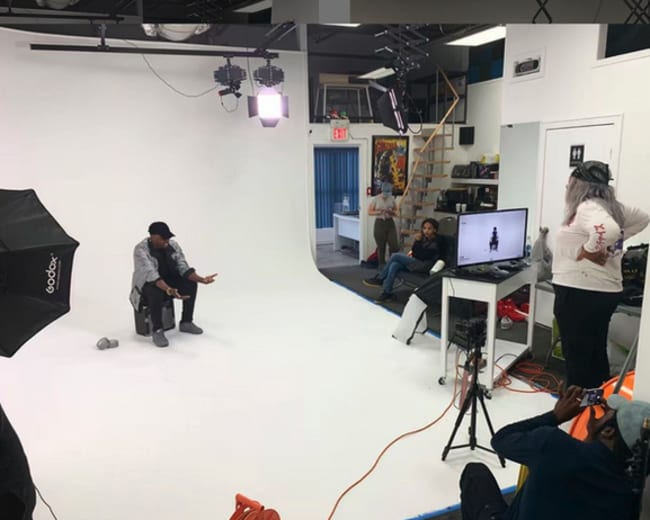 A person sits on a stool in a well-lit photography studio while others observe and operate equipment nearby.