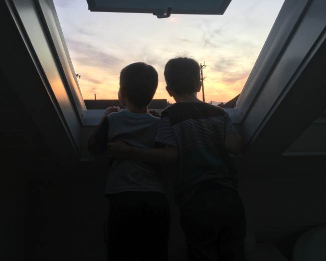 Two boys stand side by side, looking out of a skylight at a colorful sunset.