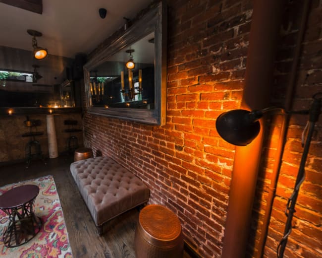 The image shows a cozy bar area with exposed brick walls, low lighting, and a mix of seating, including a tufted sofa and wooden stools.