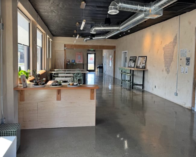 The image shows an interior space with a wooden counter, polished concrete flooring, and a minimalist design featuring large windows and industrial-style ductwork.