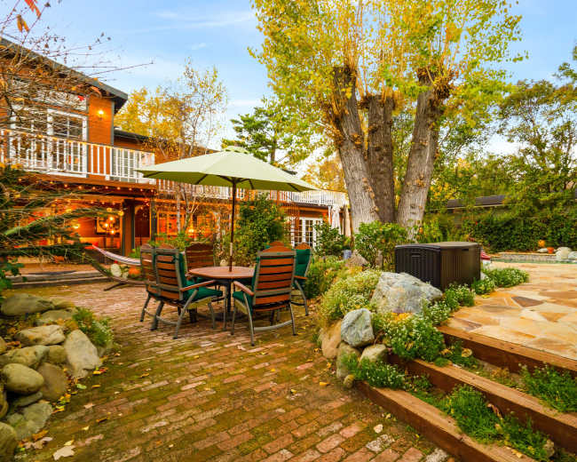 A brick patio area with a table and chairs, a green umbrella, a large tree in the background, and a wooden structure nearby.
