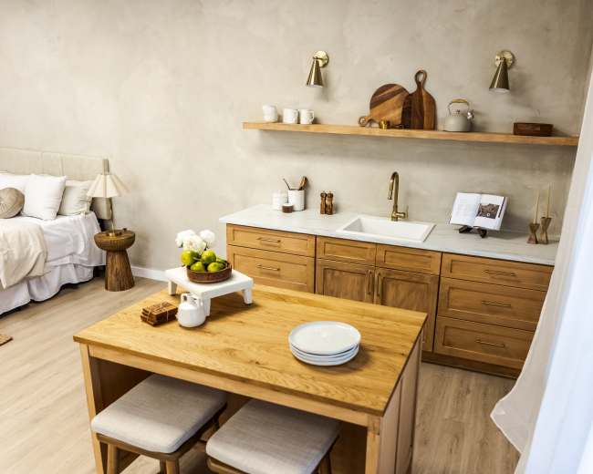 A modern kitchen with wooden cabinetry, a white sink, and a small dining table set with plates, alongside a bed and decorative items in a softly lit room.