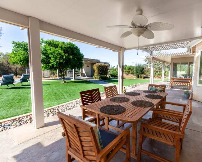 The image shows a covered patio with a wooden dining table and chairs, overlooking a landscaped yard with a green lawn and trees.
