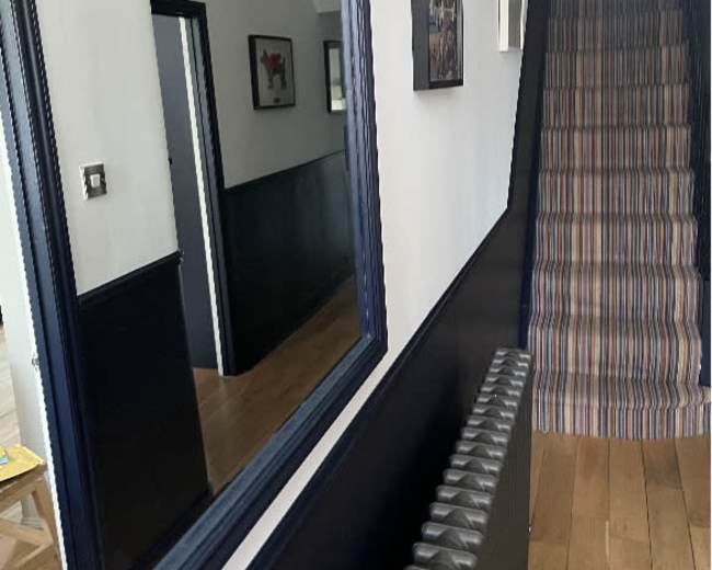 A hallway with a staircase leading upward, featuring a radiator, a mirror on the left wall, and two chandeliers hanging from the ceiling.