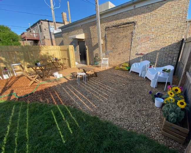 The image shows a gravel-covered outdoor space with wooden fencing, chairs, tables, and potted flowers, illuminated by sunlight and casting long shadows.
