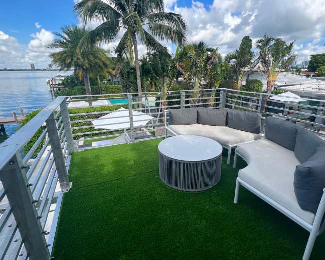 A rooftop terrace features a round table and a white sectional sofa with gray cushions, surrounded by palm trees and a view of the water.