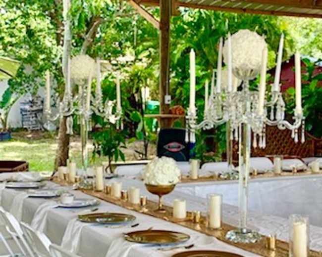 A long table is set outdoors under a wooden structure, decorated with white tablecloths, gold accents, and candelabras amidst greenery.