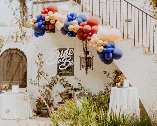 A decorated entrance features a balloon arch in blue, gold, and red, with a sign reading "Bride to Be" alongside tables set for an event.