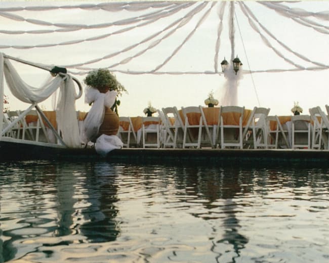 A decorated outdoor venue features a long table set with chairs under draped fabric, situated on a water-covered area.