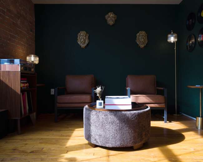The image shows a cozy living room featuring two brown leather chairs, a round coffee table, and decorative wall accents on a dark green background.