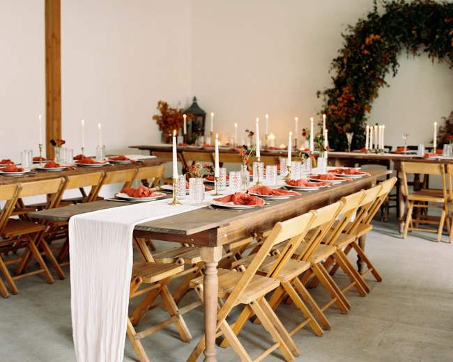 A long dining table set with plates and glassware, surrounded by wooden folding chairs, candles, and a decorative arch of foliage.