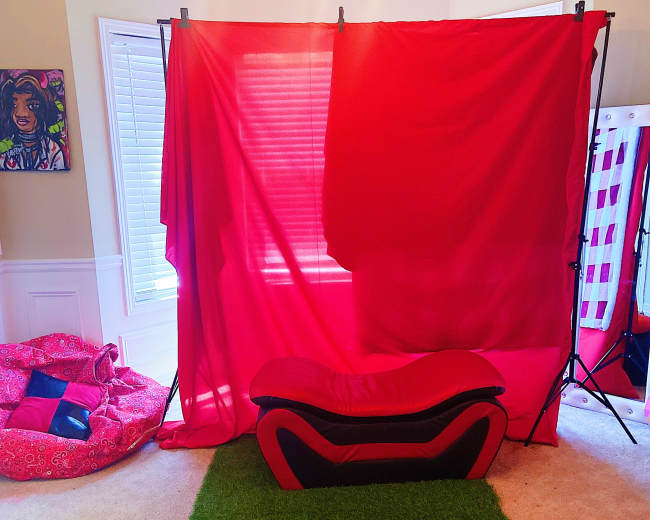 The image shows a room with a red backdrop set up, a black and red couch in front, and a green carpet on the floor.