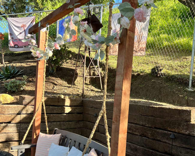 A wooden swing with cushions, adorned with flowers, placed in a grassy area near a wooden wall and hanging decorations.