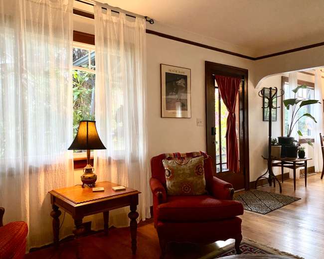The image shows a cozy living room with a lamp on a side table, a comfortable chair, sheer curtains, and a view of an entrance with a plant nearby.