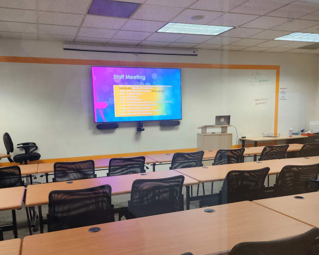 A conference room is set up for a staff meeting, featuring rows of empty chairs facing a screen displaying meeting agenda items.