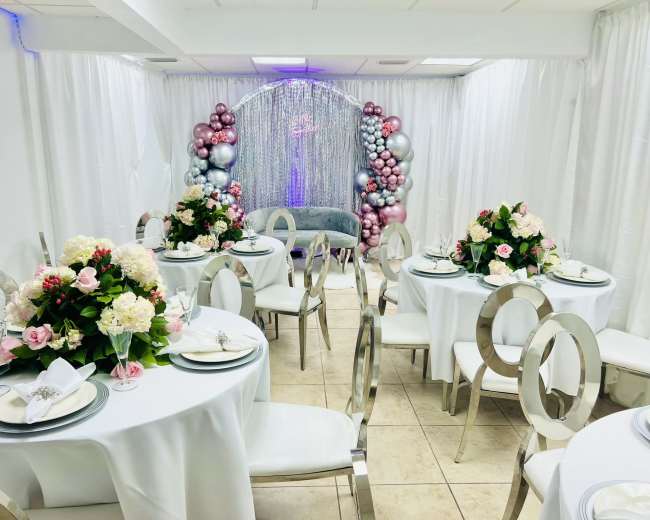 A decorated indoor venue with round tables, floral centerpieces, and a backdrop of balloons and shimmering fabric.