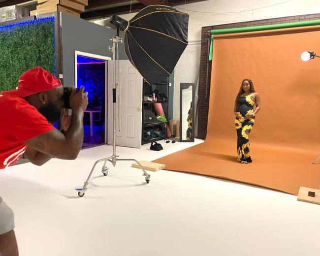 A photographer takes pictures of a model in a sunflower-patterned dress against a brown backdrop in a studio.