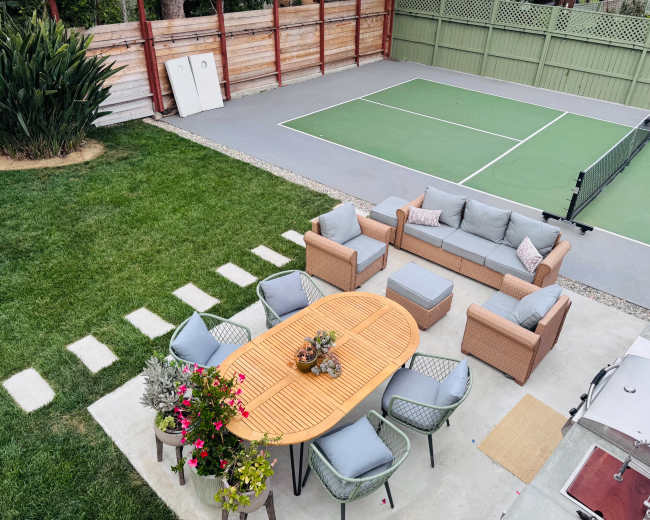 A backyard features a seating area with a round table and chairs, surrounded by green grass and a tennis court in the background.