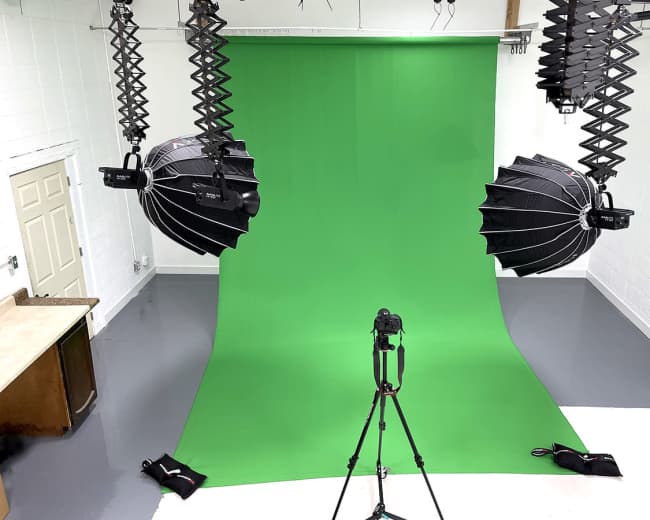 A photography studio with a green backdrop, two overhead lighting fixtures, and a camera mounted on a tripod.
