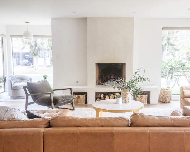 The image shows a modern living room featuring a leather sofa, a coffee table, and a fireplace, with large windows providing natural light and views of the outdoors.