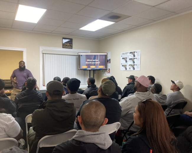 A diverse group of individuals sits in a room facing a speaker and a television displaying a presentation.