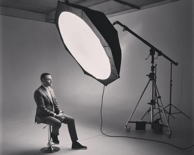 A man in a suit sits on a stool in front of a large, round softbox light in a photography studio.
