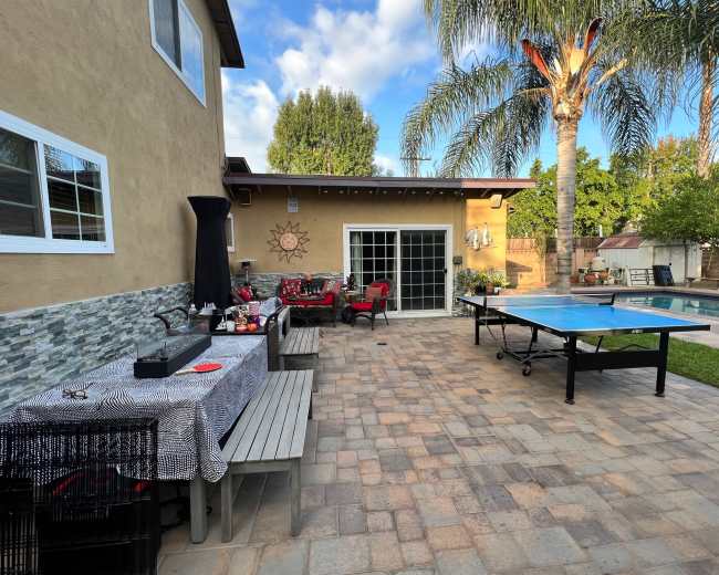 The image shows a backyard patio with a table set for dining, a ping pong table, and a swimming pool surrounded by palm trees.