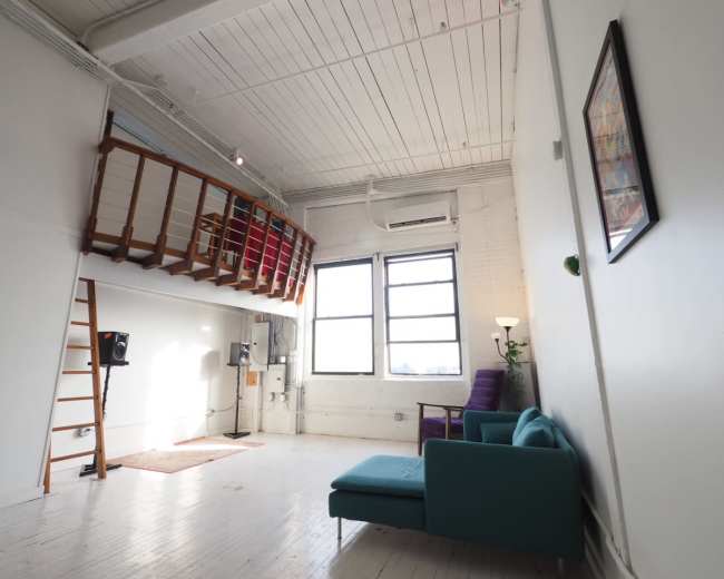 The image shows a spacious room with a high ceiling, featuring a lofted area, a sofa, a rug, and large windows allowing natural light to enter.