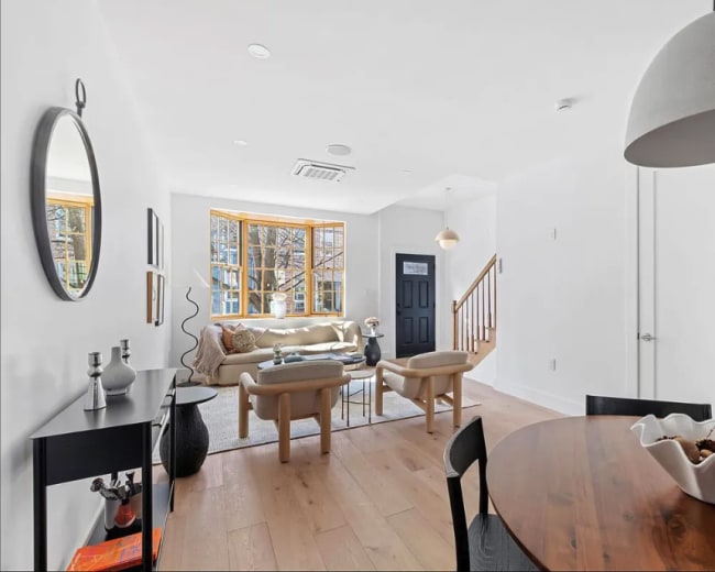 The image shows a modern living space with a beige sofa, a wooden dining table, and large windows that let in natural light.