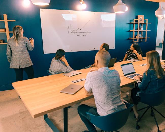A group of professionals sits around a conference table while one person presents information on a whiteboard.