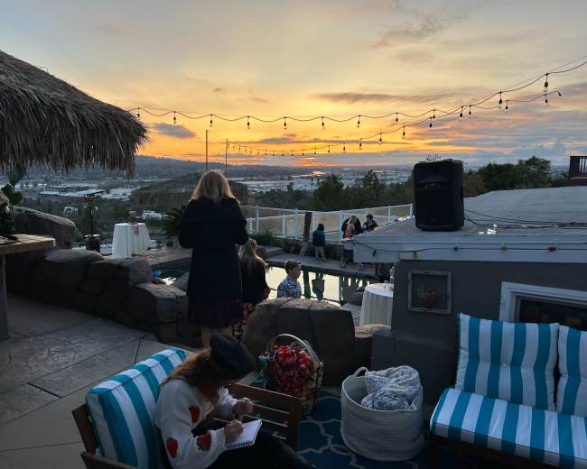 A group of people gathers on a patio overlooking a sunset, with string lights illuminated above and a pool in the foreground.