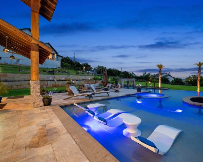 The image shows a luxurious outdoor pool area at twilight, featuring lounge chairs, palm trees, and illuminated water features.