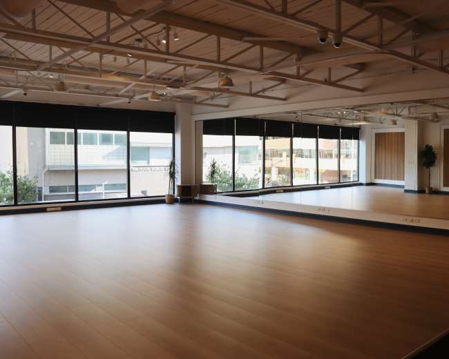 The image shows an empty, well-lit studio space with large windows and a mirrored wall, featuring wooden flooring and minimal decor.