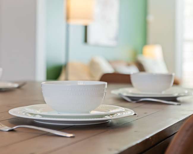 A wooden dining table is set with white plates and bowls, accompanied by stainless steel forks, in a well-lit room featuring a green wall and soft furnishings in the background.