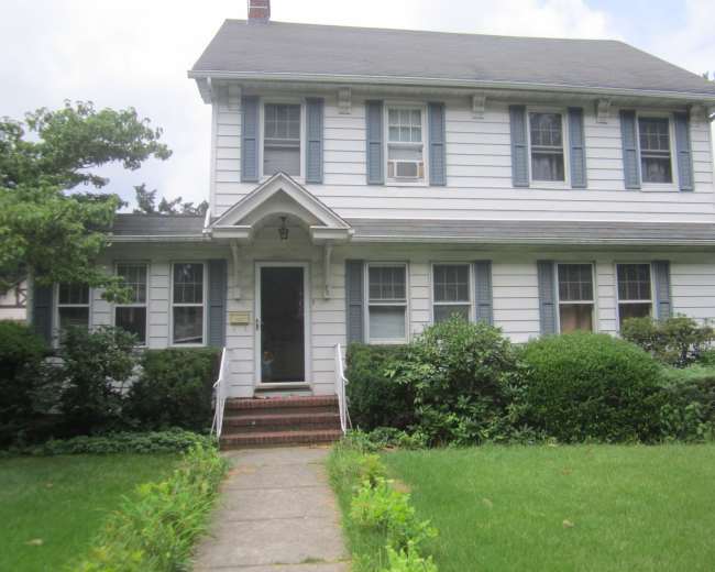 A two-story, white clapboard house with blue shutters and a front porch is surrounded by neatly trimmed bushes and grass.
