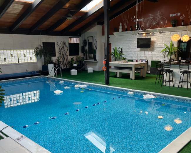 The image shows an indoor swimming pool surrounded by green artificial grass and modern furnishings, with large windows allowing natural light to enter.