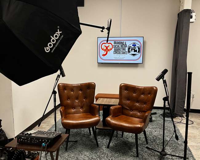 The image shows a small studio setup with two brown chairs, a microphone, and a large screen displaying branding-related content on the wall.