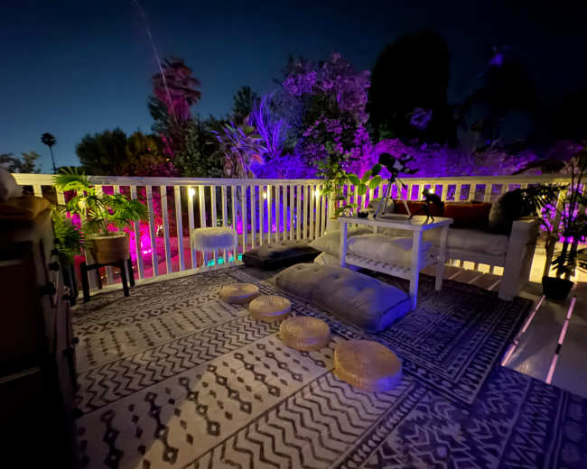 The image depicts a cozy outdoor terrace with seating, decorative cushions, and vibrant purple lighting illuminating surrounding plants at dusk.