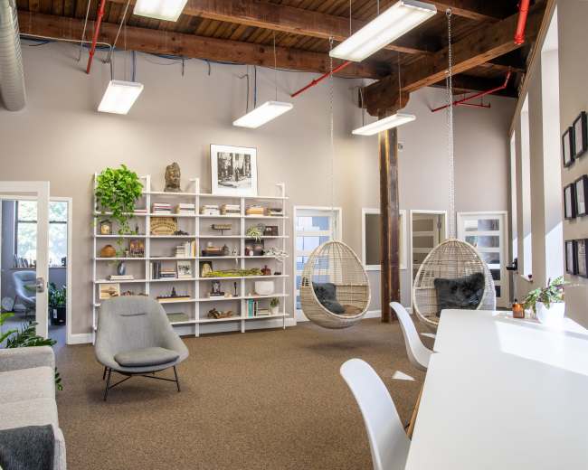 The image shows a modern office space featuring a seating area with hanging chairs, a bookshelf filled with decorative items, and large windows that provide natural light.