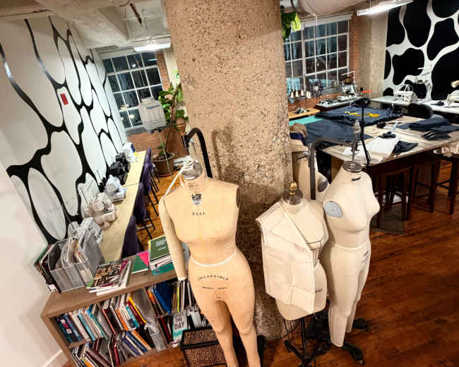 The image shows a design studio with a concrete pillar, mannequins, and a workspace filled with fabric and design materials.
