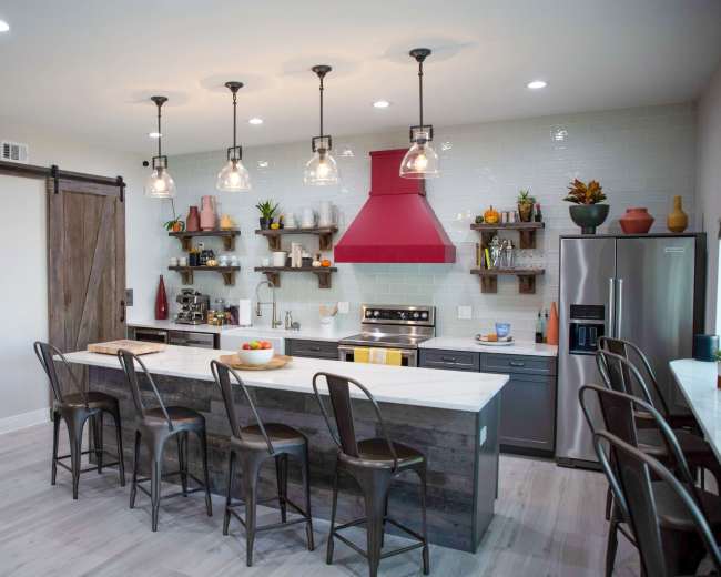 The kitchen features a large island with bar seating, stainless steel appliances, a red hood above the stove, and open shelves displaying various dishes and plants.