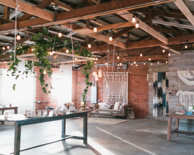 The image shows a spacious, rustic interior with wooden beams, string lights, and various decorative elements, including plants and textile art.