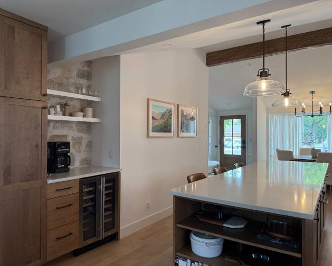 The image shows a modern kitchen featuring a wooden cabinet with a built-in wine cooler, a stone accent wall, and an island with bar seating.