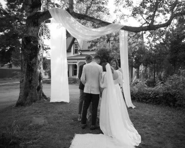 A couple stands under a draped fabric arch while exchanging vows in a garden setting.