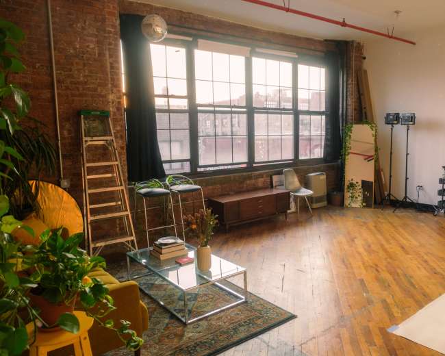 The image depicts a spacious, industrial-style studio with large windows, wooden floors, and various furnishings, including a glass coffee table, bar stools, and a desk with lighting equipment.
