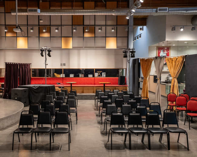 The image shows an indoor performance space set up with rows of black chairs facing a small stage, flanked by red curtains and lighting equipment.