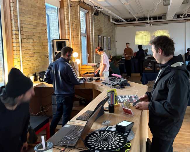 A group of people are gathered inside a well-lit room with wooden floors, engaging with music equipment and DJing setup on a long wooden table.