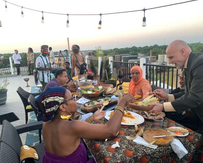 A diverse group of people shares a meal at a rooftop gathering, with food laid out on a table and city views in the background.