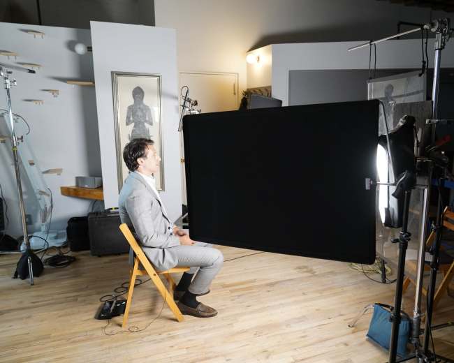 A man in a suit sits on a chair in a photography studio, facing a large black screen and surrounded by cameras and lighting equipment.